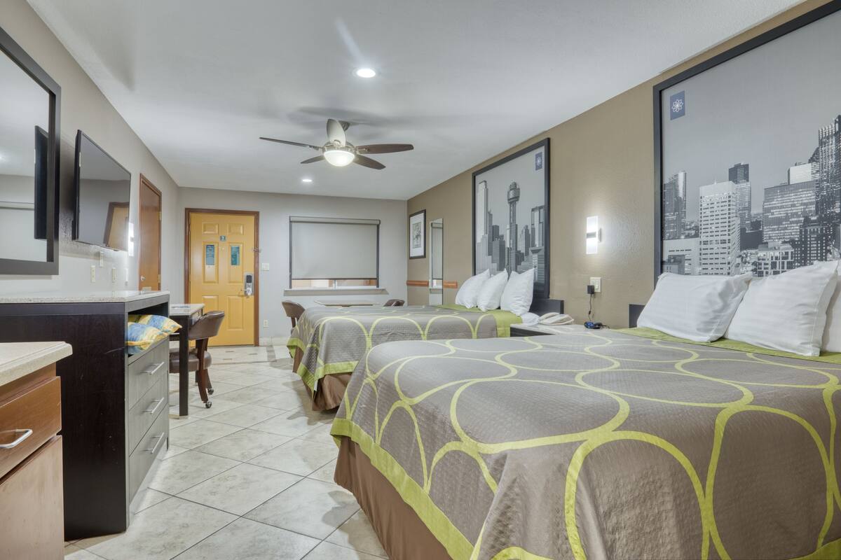 A hotel room with two beds, a desk, a chair, a wardrobe, a ceiling fan, and a cityscape poster on the wall.