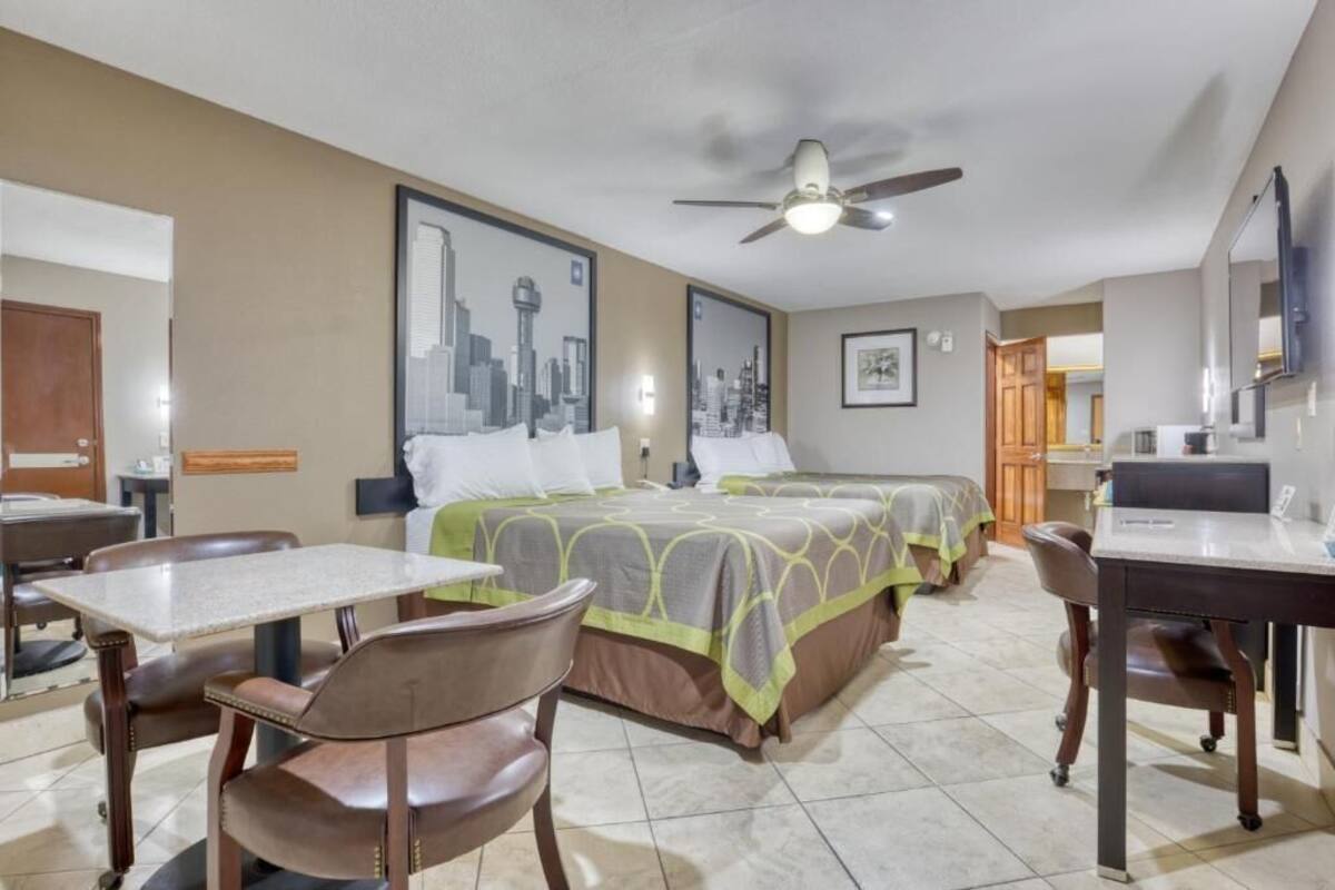 A hotel room with a bed, two chairs, a desk, a ceiling fan, and a cityscape painting on the wall.