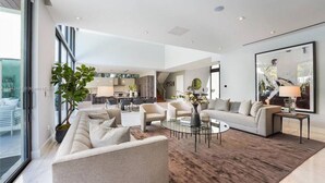 A spacious living room with a large window, a round coffee table, and a modern kitchen in the background. - Playa Villa - Modern Luxury Redefined (Miami Beach)