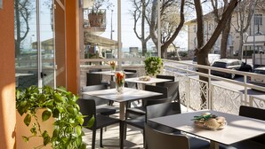 Un café al aire libre con mesas y sillas, plantas en macetas y vista a la calle.