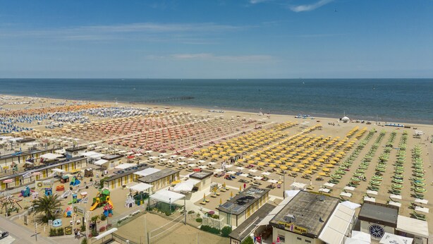 Una playa con hileras de sombrillas y tumbonas, un parque infantil y un bar.