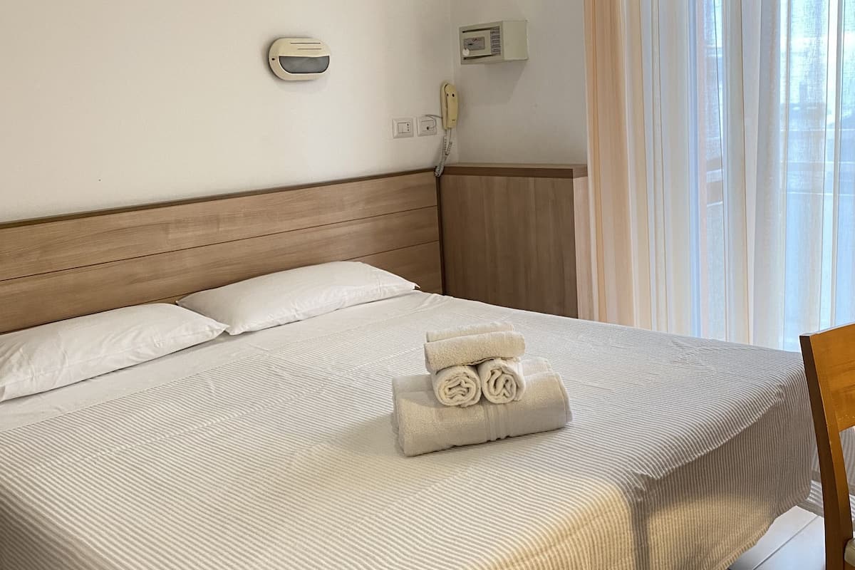 A neatly made bed with a striped bedspread, a wooden headboard, a bedside telephone, and a window with sheer curtains.