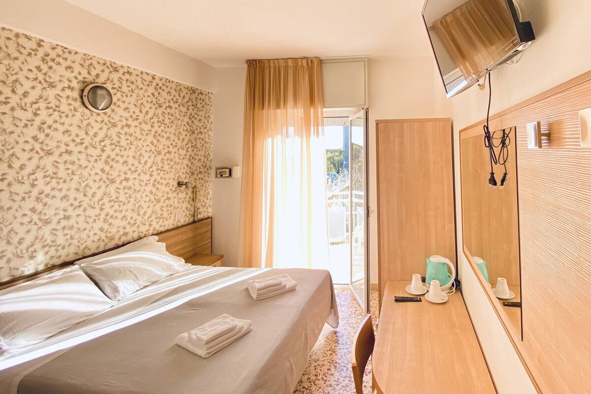 A hotel room with a bed, a desk, a mirror, and a view of the outdoors.