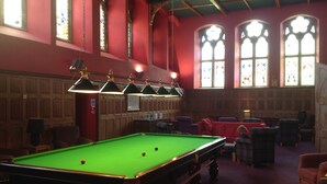 Une salle de billard équipée d’une table de billard, de chaises et d’un mur rouge orné de vitraux.