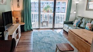 A cozy living area with a sofa, a TV, a wooden table, and a balcony with a view of the city.