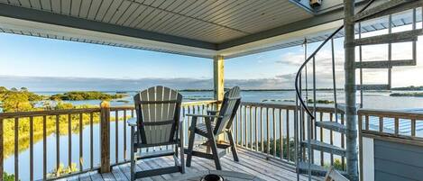 Une terrasse en bois avec des chaises Adirondack et une table, offrant une vue sur une étendue d’eau et la verdure environnante.