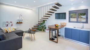 Una sala de estar moderna con un sofá gris, una mesita redonda y una escalera que lleva a una zona de cocina con armarios azules y una ventana.