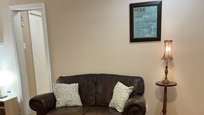 A living room with a brown leather couch, a glass coffee table, a wooden floor, a framed quote on the wall, and a lamp on a wooden stand. - Private, Quiet Ap. “Morning Singing Birds”, close to Phoebe (Albany)