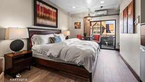 A bedroom with a large bed, two bedside tables, a chandelier, and a view of the outside.