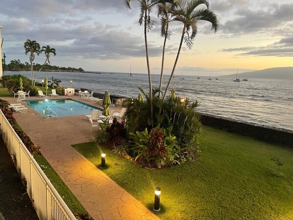 A pool area with lounge chairs and umbrellas, a well-maintained lawn, and a view of the sea with boats.