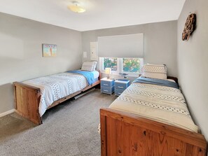 Une chambre avec deux lits en bois, une fenêtre avec des stores, une table de chevet avec une lampe et un petit tableau décoratif au mur.