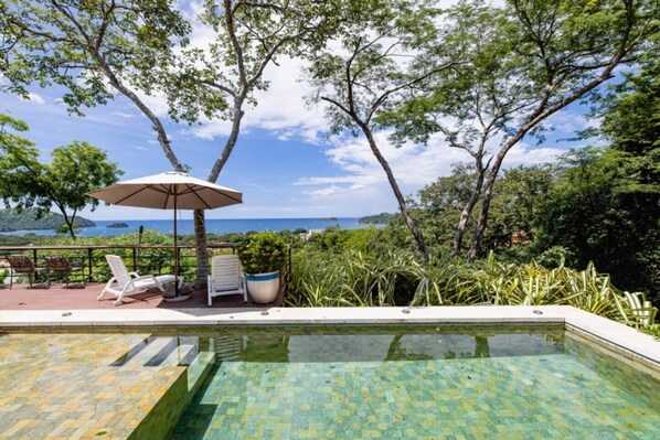 Zona de piscina con sillas de descanso y una sombrilla, rodeada de vegetación exuberante y vistas al mar.