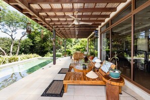 Un área de estar al aire libre techada con piscina, mobiliario de madera y plantas en macetas.