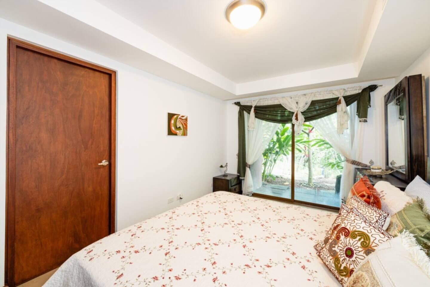 Une chambre avec un grand lit, une porte en bois, une table de chevet, une commode, une fenêtre avec des rideaux et une vue sur la verdure à l’extérieur.
