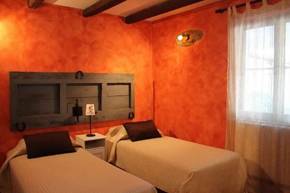A room with two beds, a wooden headboard, a small bedside table with a lamp, and a window with curtains.