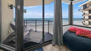 Un balcón con vistas a la playa y al océano, una cama con almohadones rojos y una pequeña zona de estar con una mesa y sillas.