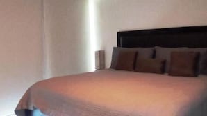 Una cama bien hecha con cabecera oscura, una lámpara de noche y una pared lisa.
