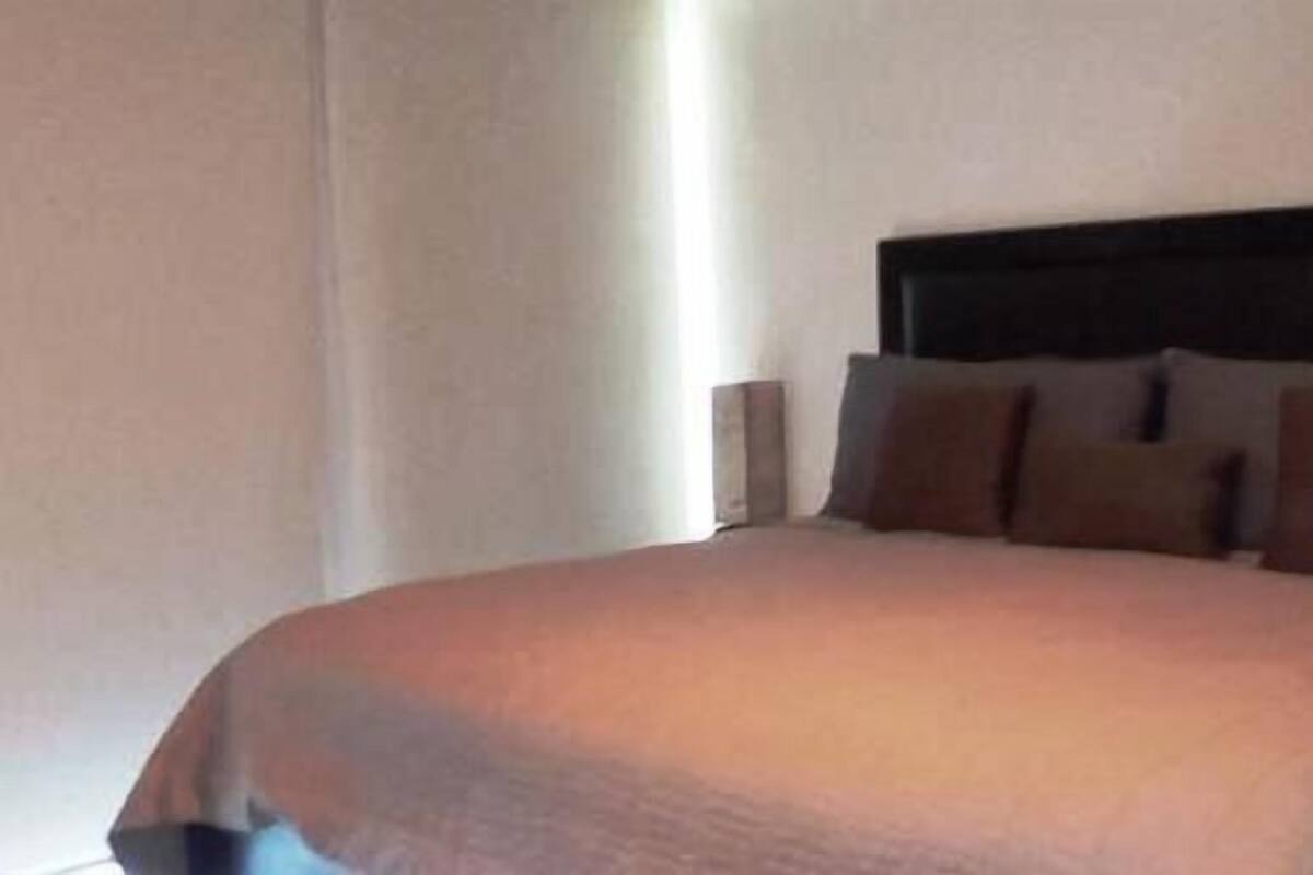 A neatly made bed with a dark headboard, a bedside lamp, and a plain wall.