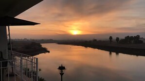 Vista del atardecer desde un balcón con vistas a un río tranquilo, con un poste de luz y barandilla a la vista.