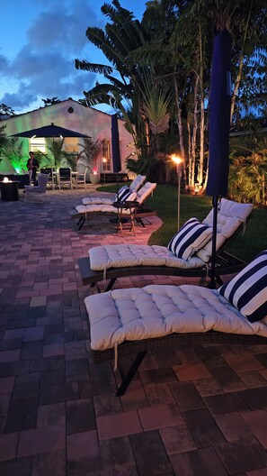 Outdoor lounge chairs with cushions, a paved patio, and a building with a lighted entrance. - Capri Cottage, Pool, walk to restaurants! (West Palm Beach)