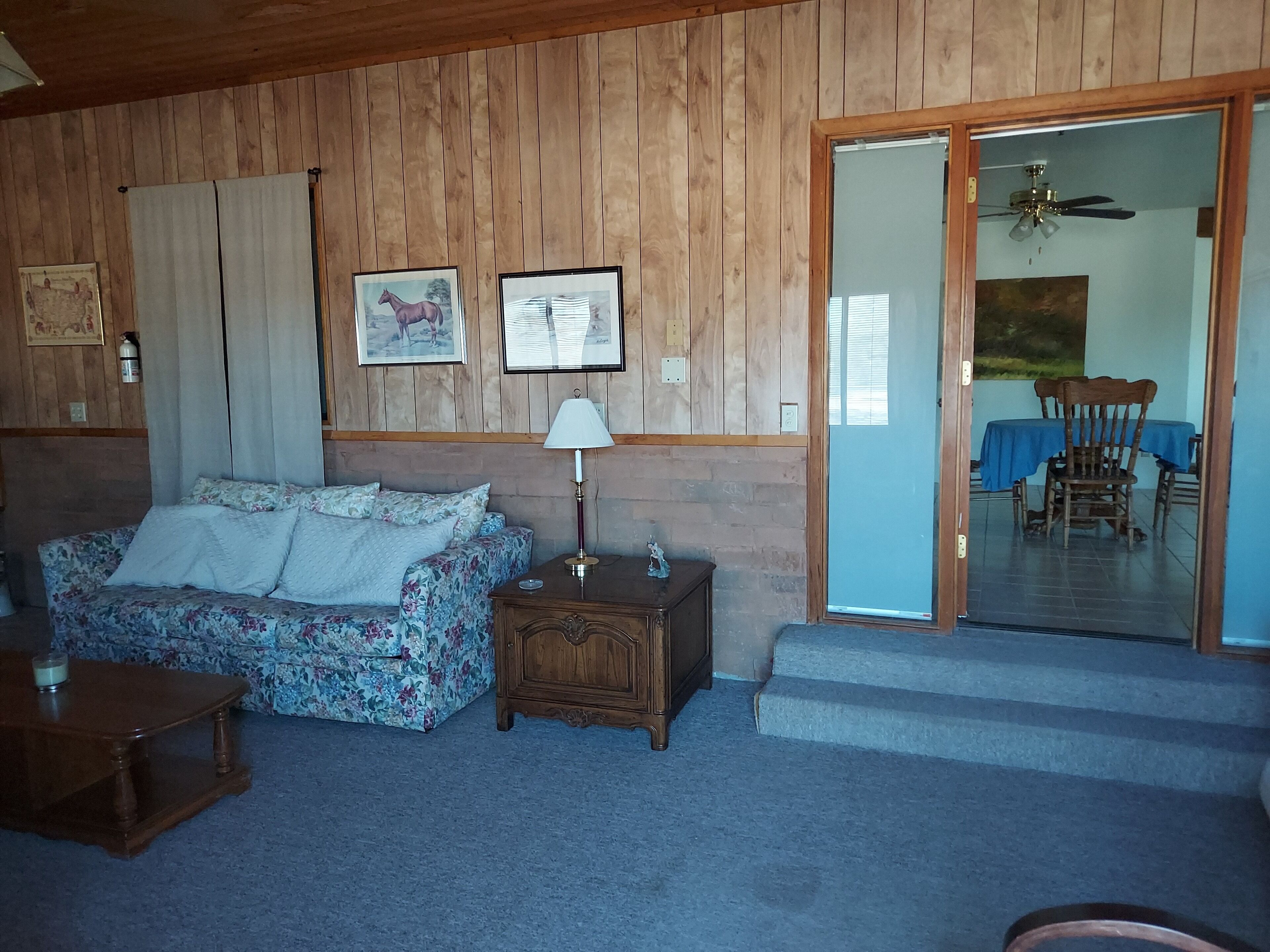 A cozy room with a floral sofa, a small wooden nightstand with a lamp, and a dining area with a table and chairs.