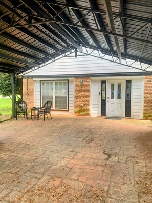 A covered patio area with a brick floor, a table with chairs, and a building with a brick facade and large windows. - Cozy Private Room-Queen&Full Bed, Hot Tub Pool Pets Welcome! (Milton)