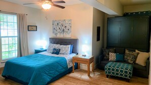 A bedroom with a bed, nightstands, a ceiling fan, a window with curtains, a dark wardrobe, and a patterned ottoman.