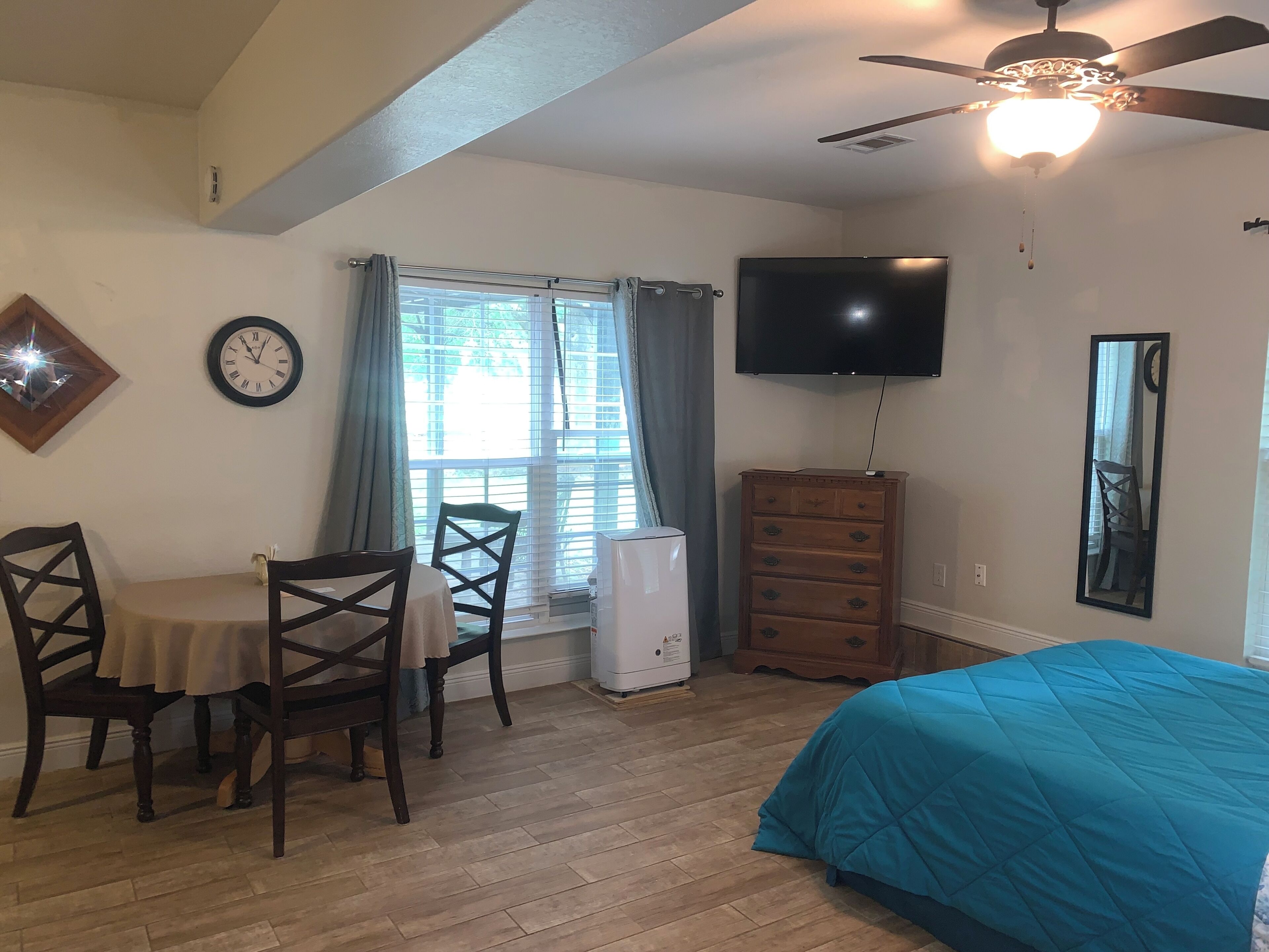 Un dormitorio con cama, cómoda, televisor, ventilador de techo, ventana con cortinas y una mesa de comedor con sillas.