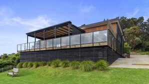 Una casa moderna con un amplio balcón de vidrio, terraza de madera y un patio cubierto de césped.