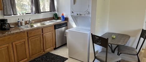 Una cocina compacta con gabinetes de madera, lavavajillas integrado, refrigerador blanco y un comedor con dos sillas y una mesa.