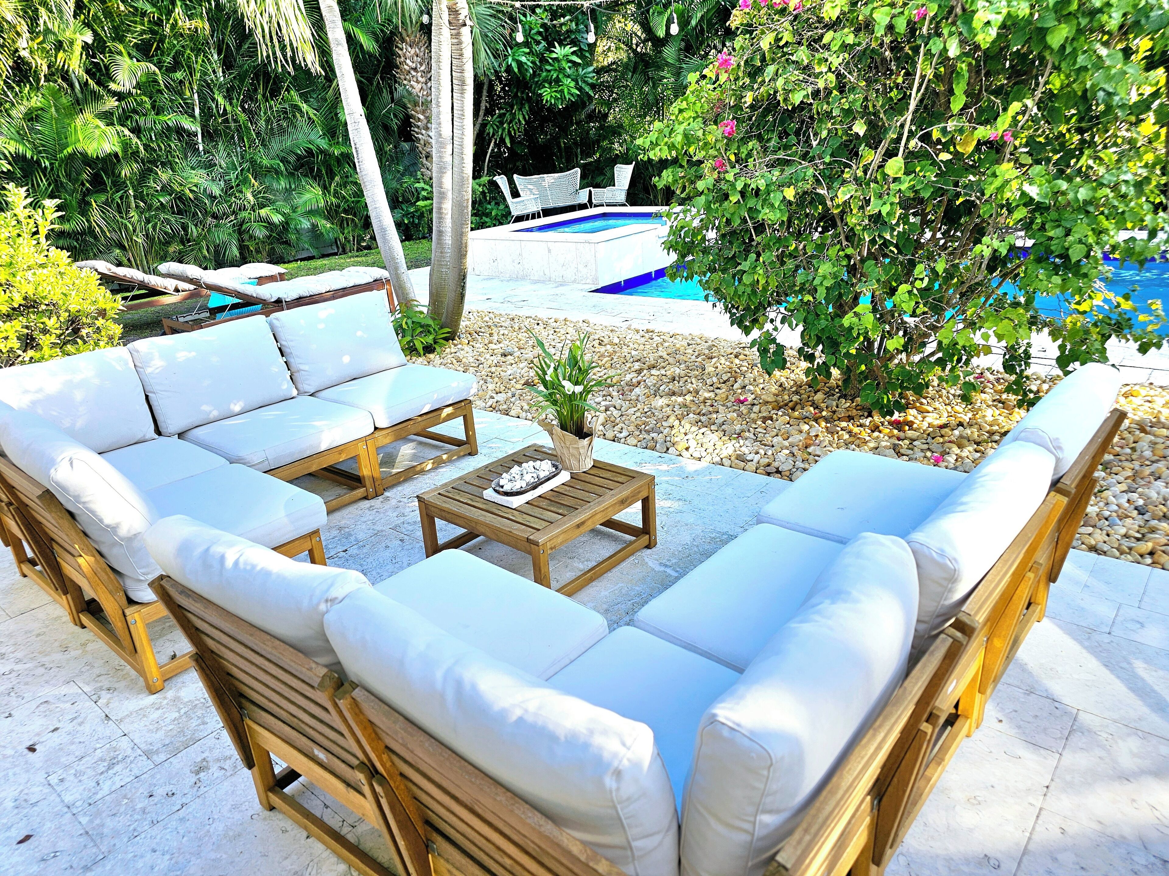 A sunlit outdoor seating area with a pool, surrounded by lush greenery and a pebble path.