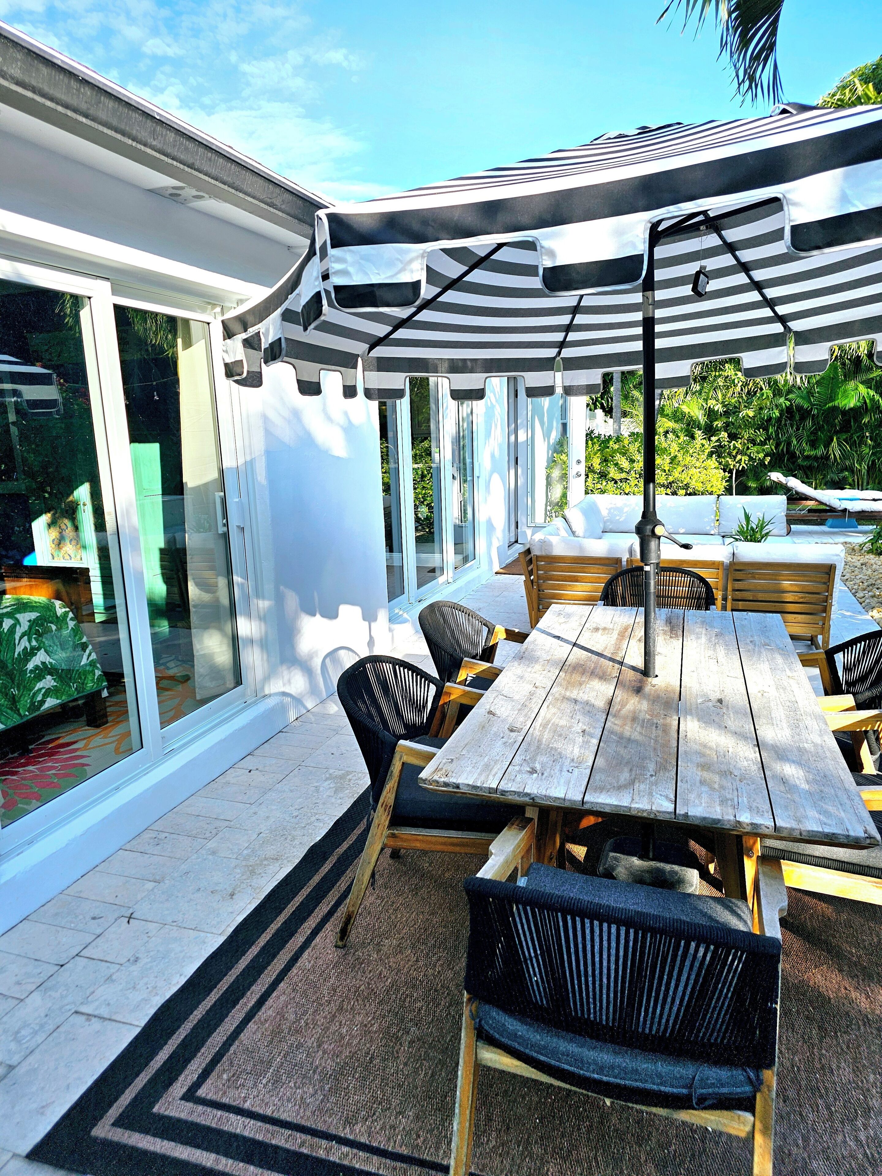Un patio al aire libre con mesa y sillas de madera, un toldo a rayas y una puerta de vidrio que da a un jardín.