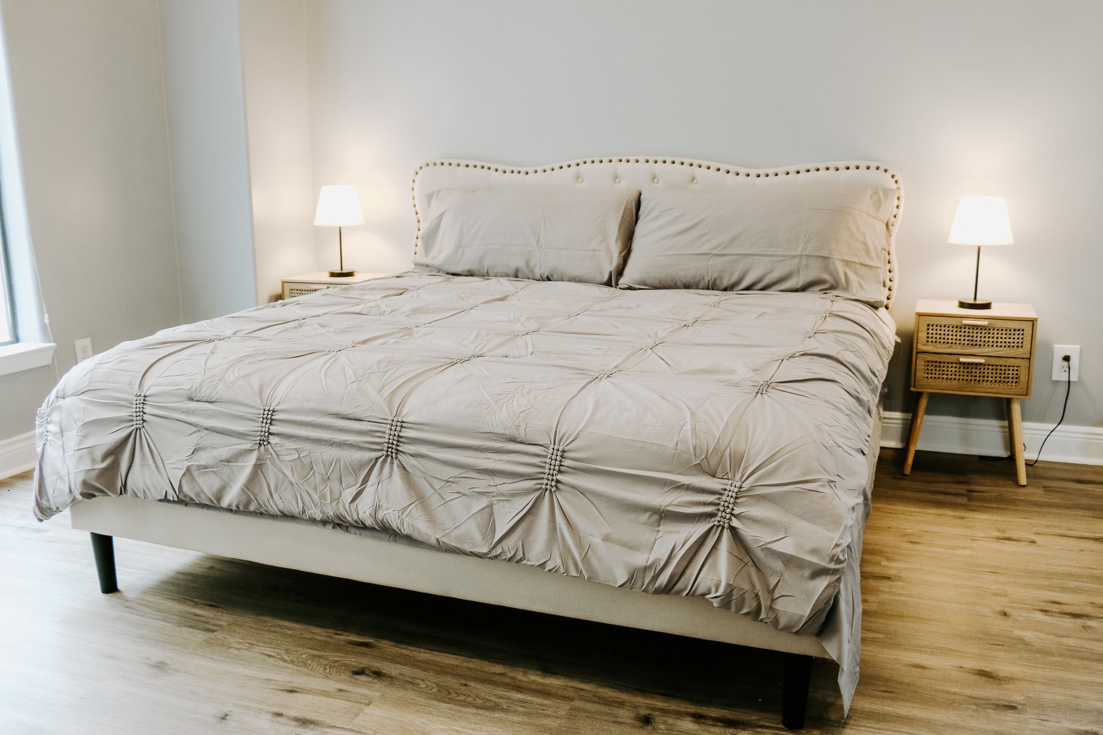 Una cama bien hecha con cabecera capitonada, dos mesitas de noche con lámparas y suelo de madera.