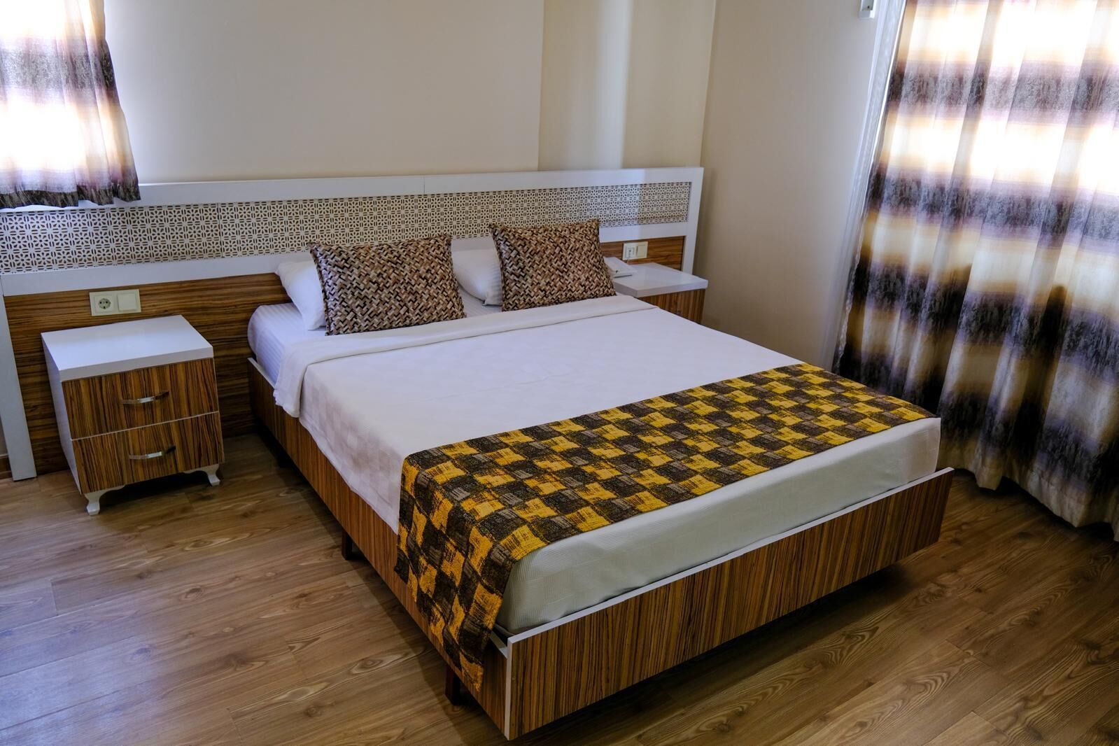 A hotel room with a large bed, wooden headboard, two bedside tables, and a patterned throw blanket.