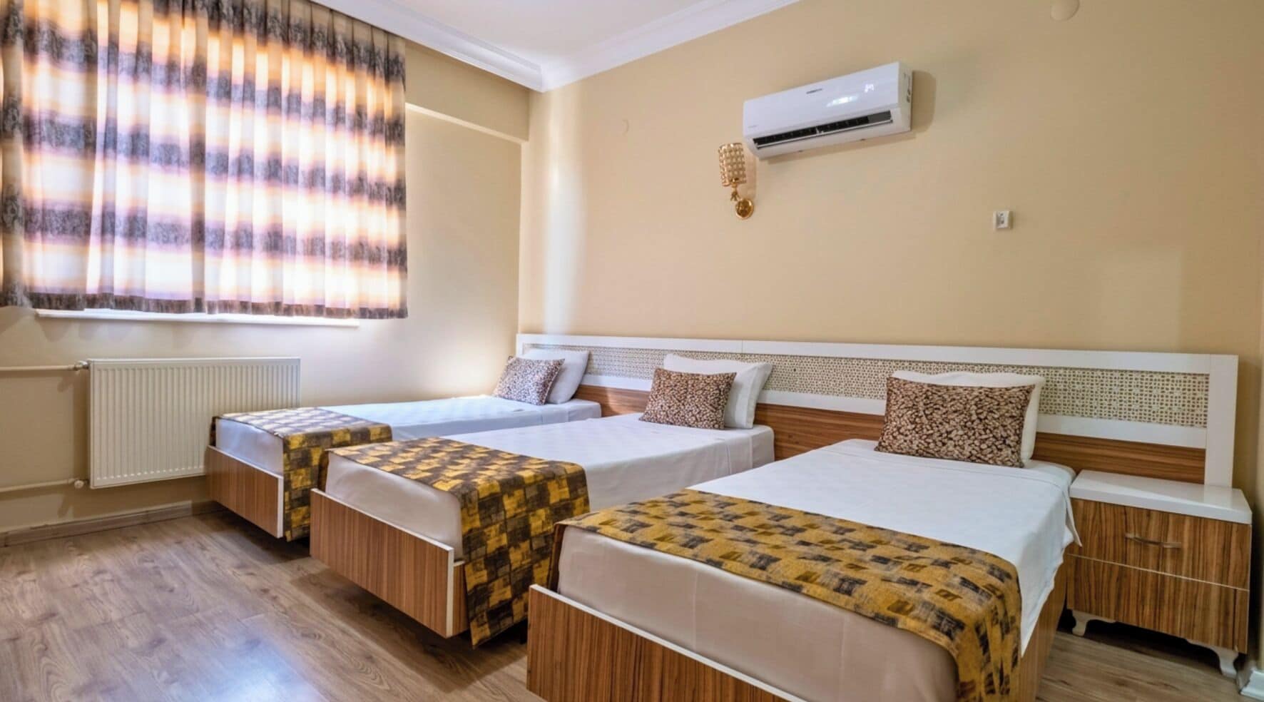 A hotel room with two single beds, a radiator, an air conditioning unit, and a window with curtains.