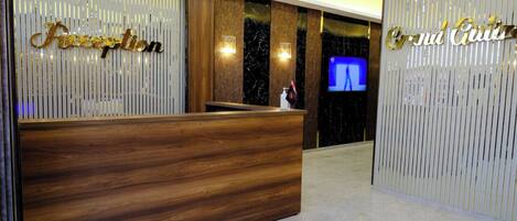 A reception desk with a wooden finish, flanked by vertical panels with the words 'Accents' and 'Grand Cuts' in gold lettering. A television is mounted on the wall to the right.