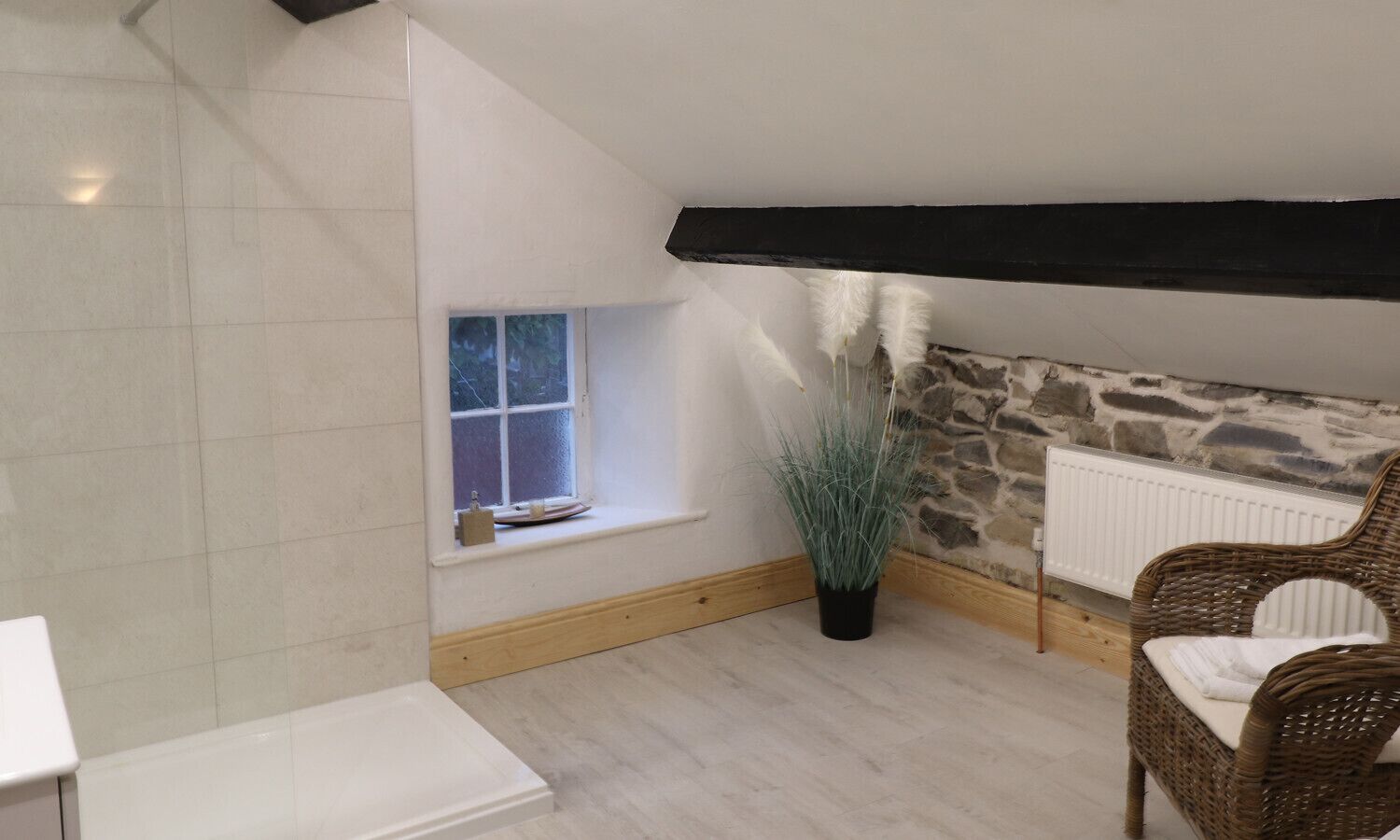 Una habitación pequeña y acogedora con techo inclinado, ventana con vistas, pared de piedra, radiador y una silla de mimbre.