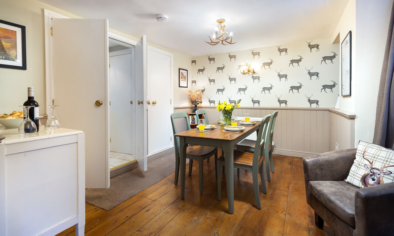A dining area with a wooden table set for four, surrounded by chairs. A chandelier hangs from the ceiling, and the walls are decorated with a pattern of deer silhouettes.