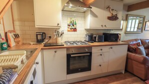 A compact kitchen with a gas stove, oven, and microwave. There is a wooden countertop, a sink, and a dining area with a brown leather couch. - Heron Cottage (Ambleside)