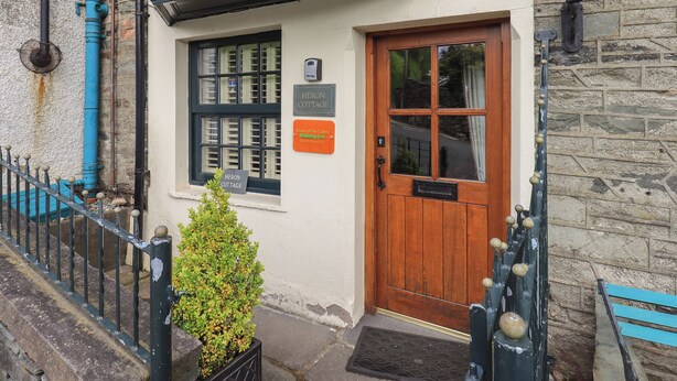 A cottage with a wooden door, a window with shutters, and a sign that reads "Heron Cottage." - Heron Cottage (Ambleside)