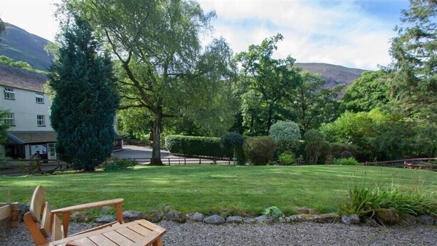 A wooden deck with chairs, a well-maintained lawn, and a variety of trees and shrubs. - Swinside Cottage (Keswick)