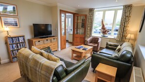 A living room with a television, bookshelves, a sofa, a coffee table, and a window with curtains. - Middlerigg (Windermere)