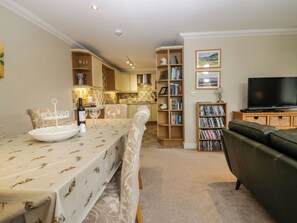 A dining area with a table set for two, a bookshelf with books, a television, and a kitchen in the background. - Middlerigg (Windermere)