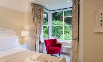 A hotel room with a bed, a red chair, a window with a view of greenery, and a framed picture on the wall.