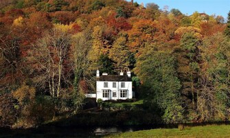 A white house with multiple windows, surrounded by dense trees in autumn colors, and a body of water in the foreground.