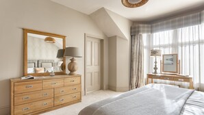 A bedroom with a large bed, a wooden dresser, a mirror, and a bedside table with a lamp and picture. - Archie's (Ambleside)