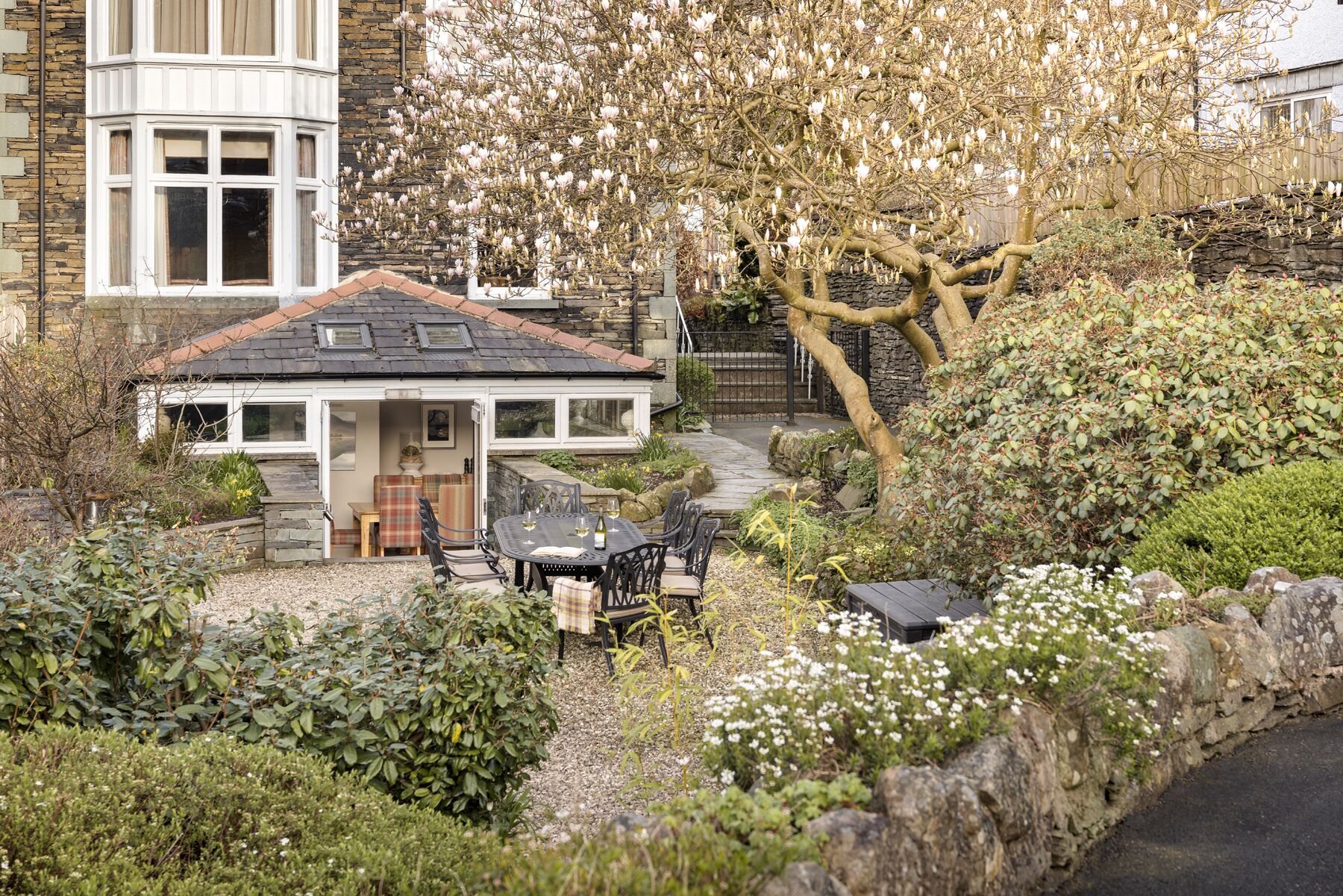 Una casa con fachada de piedra, un porche cubierto con mobiliario exterior y un jardín con diversas plantas y flores.