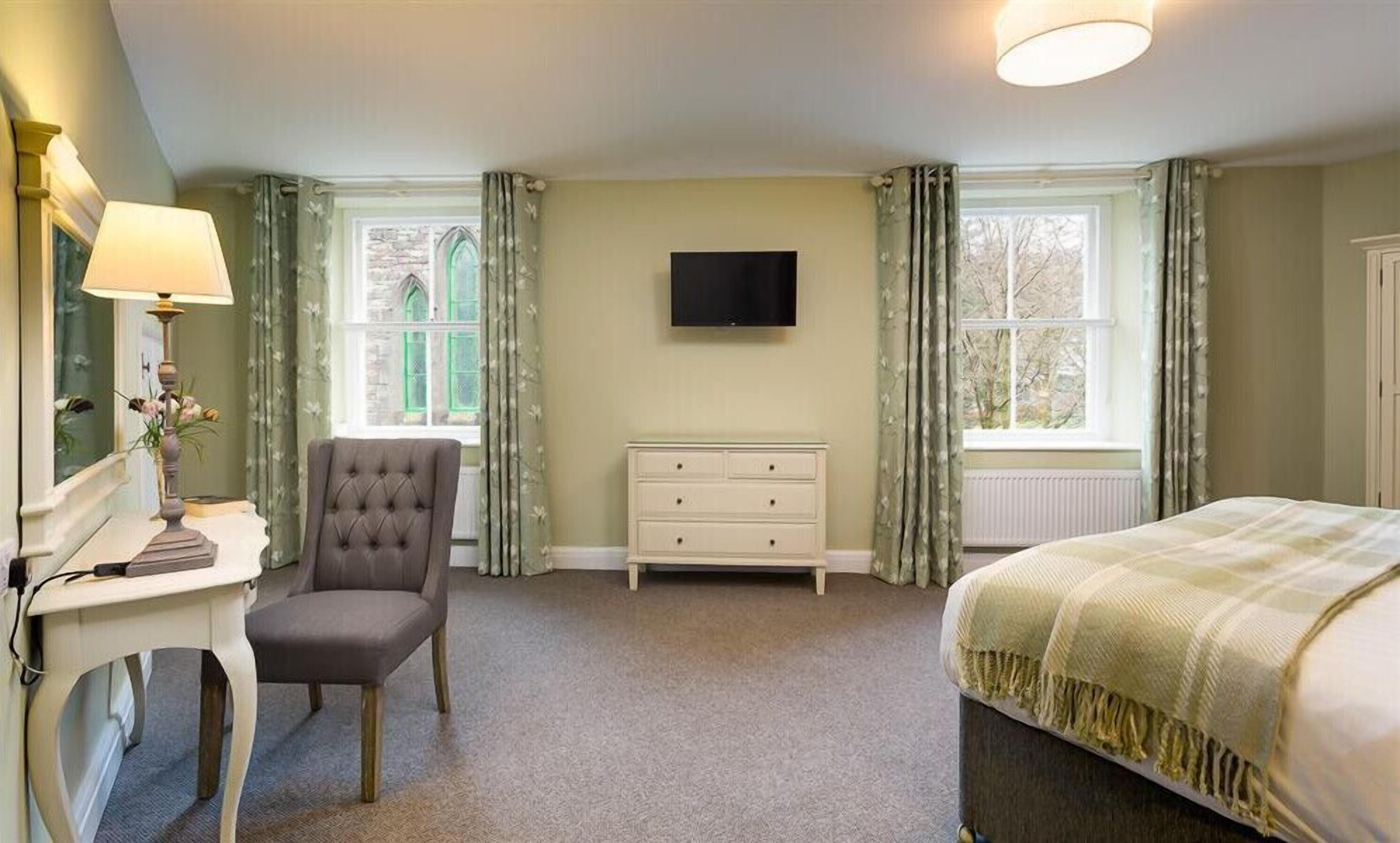 A bedroom with a bed, a desk with a lamp, a chair, a dresser, and a TV on the wall.