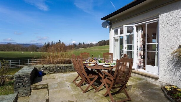 A patio with a table set for dining, stone flooring, and a view of a green field and mountains. - Juniper Cottage (Windermere)