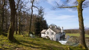 A white house with a sloped roof, surrounded by trees and a grassy hill. - Juniper Cottage (Windermere)